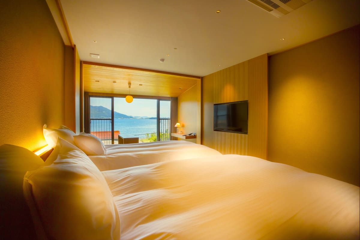 A hotel room with two large beds, a TV mounted on the wall, and a view of the sea through large windows.