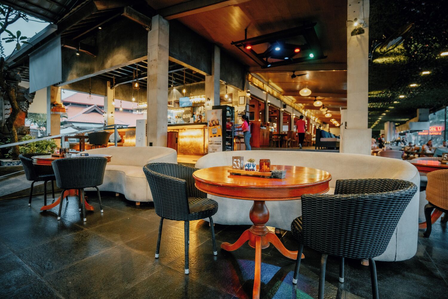 A modern indoor seating area with round tables and wicker chairs, featuring a bar in the background.