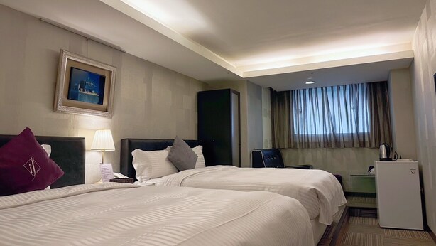Habitación de hotel con dos camas, un escritorio, una silla, un refrigerador pequeño y una ventana con cortinas.