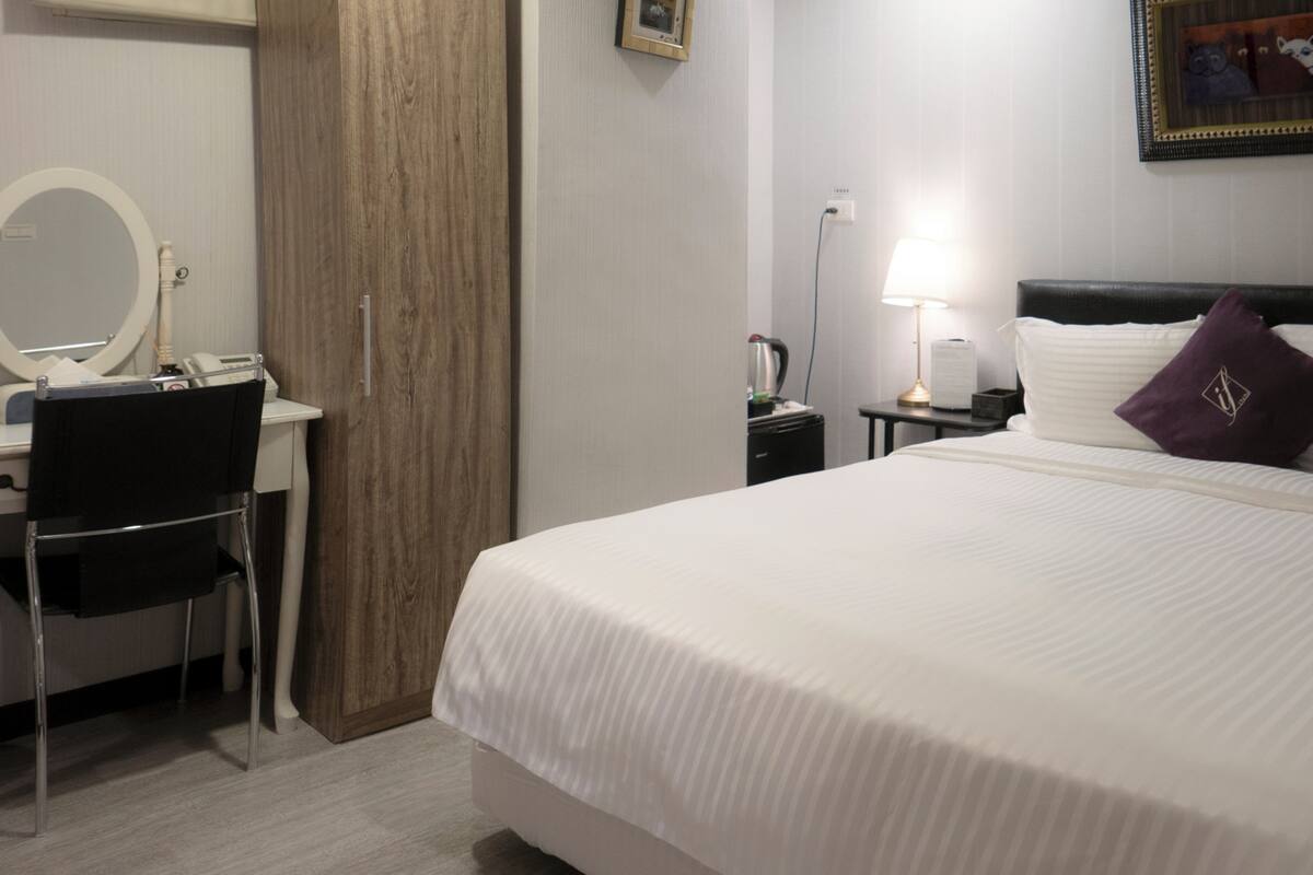 A hotel room with a bed, a desk with a chair, a bedside table with a lamp, and a wall-mounted picture.