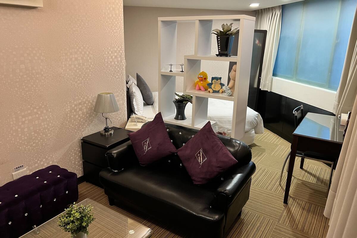 A hotel room with a black leather sofa, a small table with a plant, a TV, a bookshelf with decorative items, and a window with curtains.