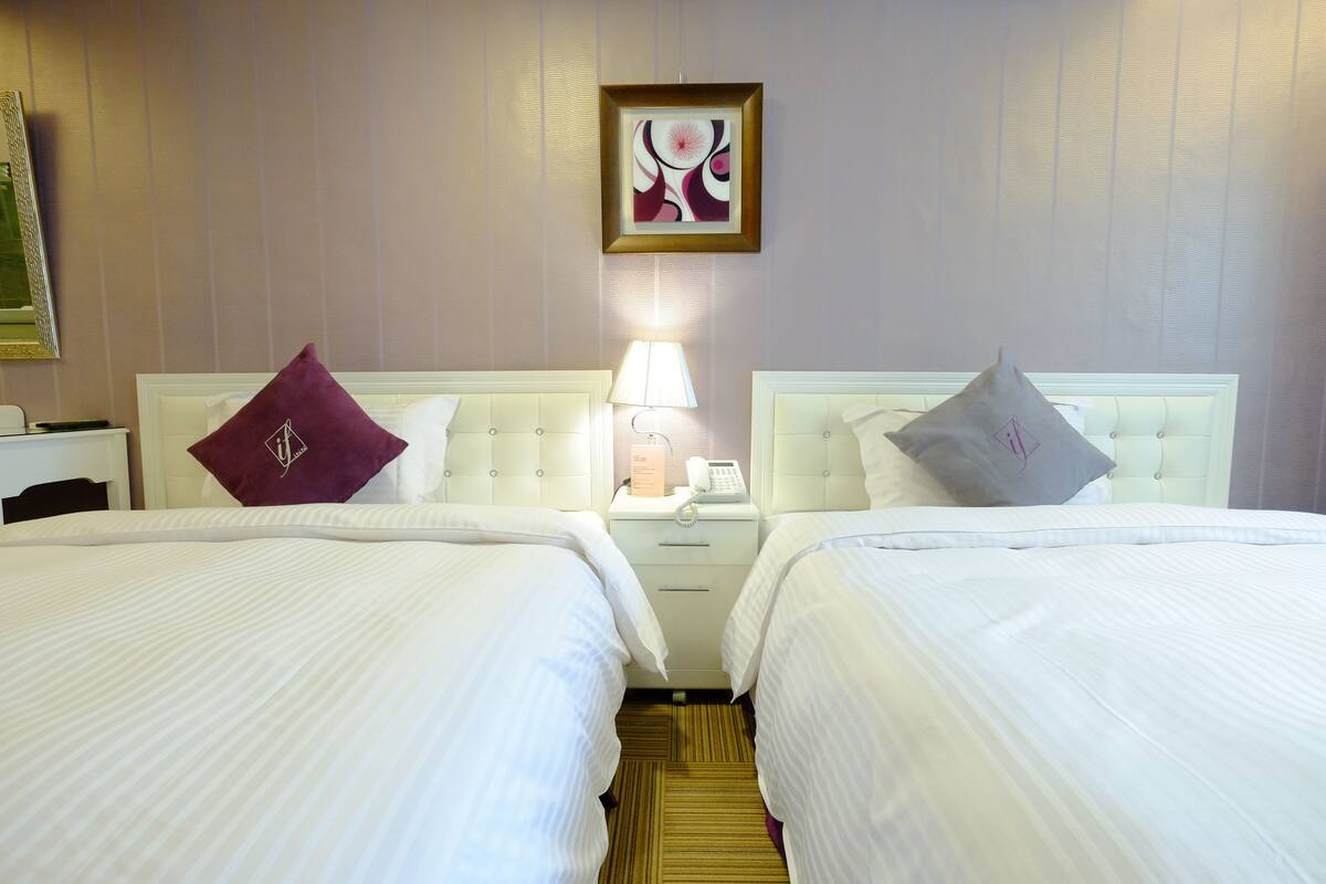 A hotel room with two beds, each with a headboard and a pillow. There is a nightstand with a lamp and a phone between the beds.