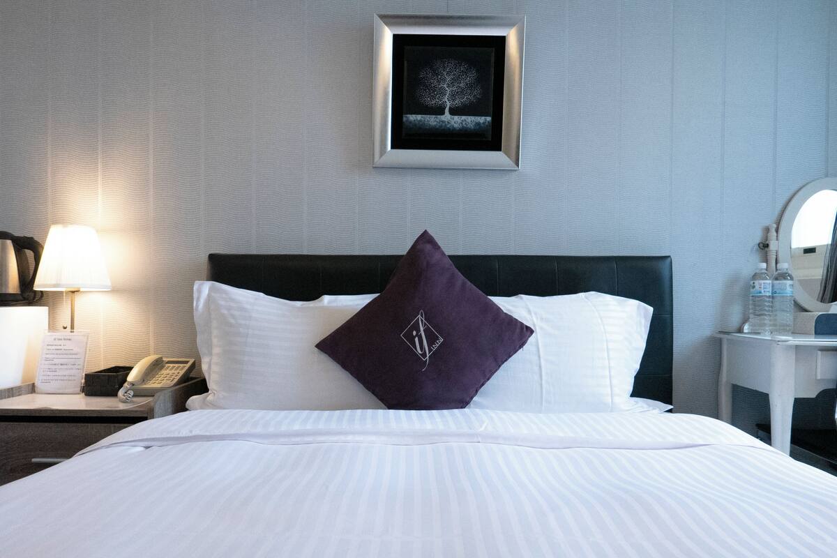A hotel room with a neatly made bed, a framed artwork above the bed, a bedside table with a lamp, a phone, and a water bottle.
