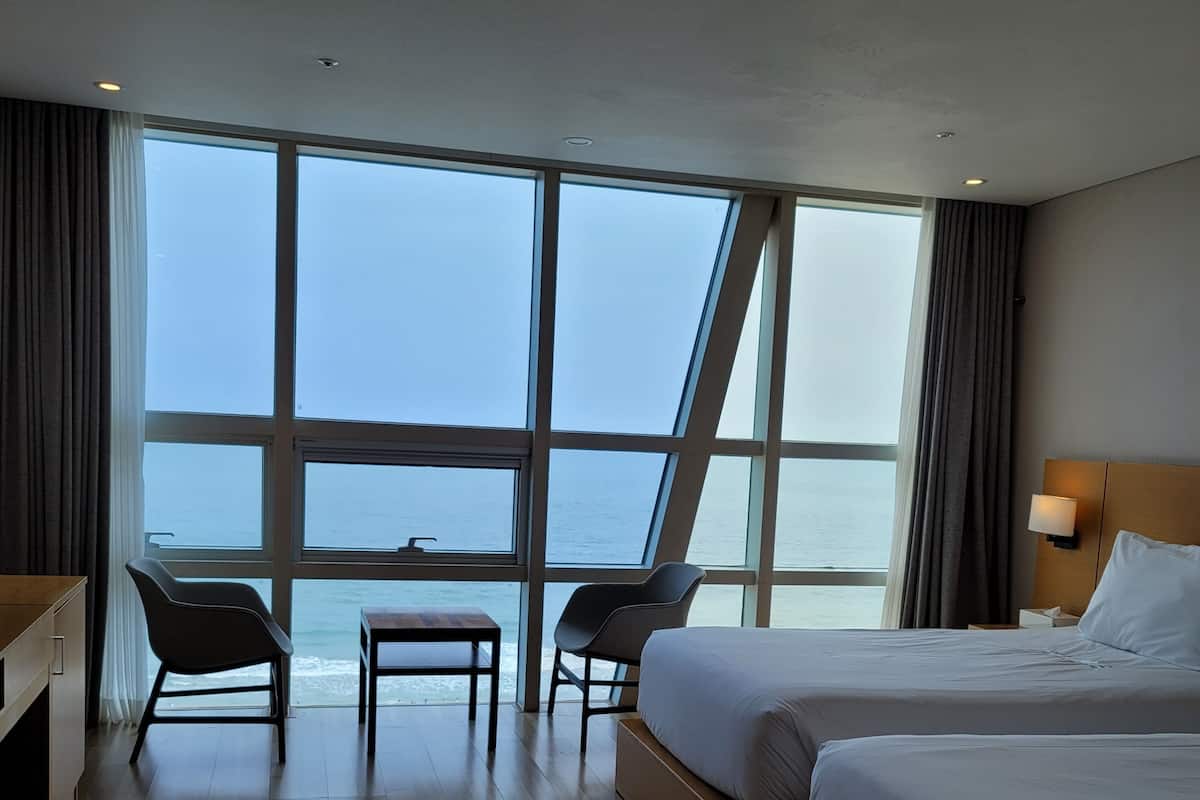 A hotel room with two beds, a desk, and a chair, large windows with a view of the sea, and a lamp on the nightstand.