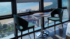 Ein Zimmer mit großen Fenstern mit Blick auf Strand und Stadtlandschaft. Zwei Stühle und ein kleiner Tisch sind vorhanden.