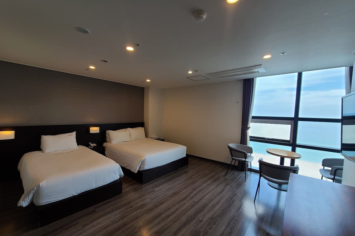 A hotel room with two beds, a small table, a chair, and a large window offering a view of the sea.