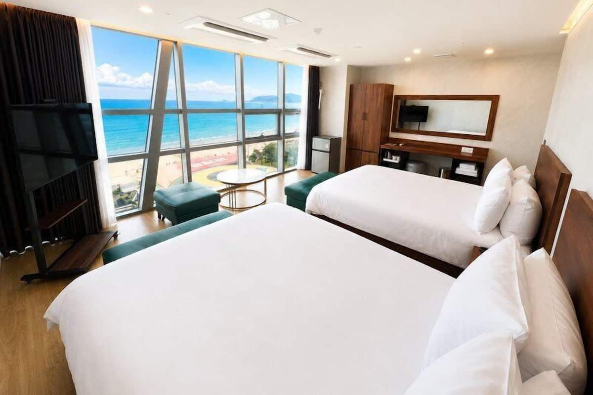 A hotel room with two beds, a TV, a desk, and a view of the beach.