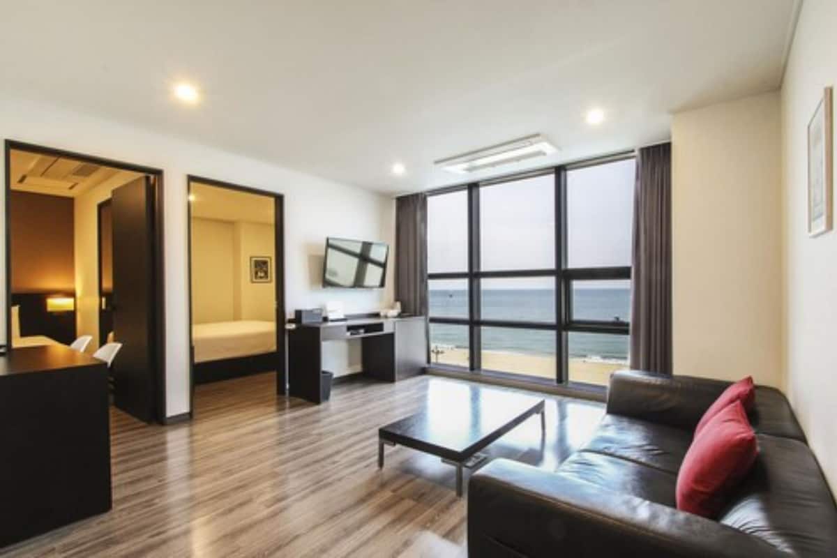 A modern hotel room with a large window offering a view of the sea, a cozy seating area, a small kitchenette, and a separate sleeping area.