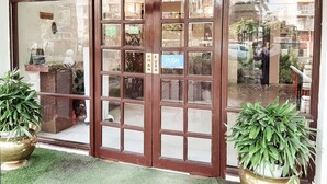 Fachada con puerta y ventanas de vidrio amplias, plantas en macetas y un cartel que indica servicio de asistencia.