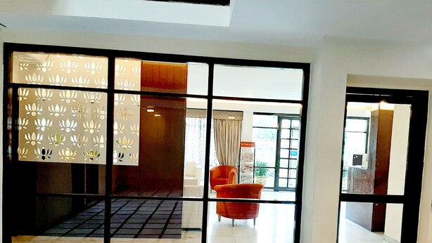 Un vestíbulo de hotel con piso de baldosas, un ventanal con celosía decorativa y un área de descanso con sillas anaranjadas.