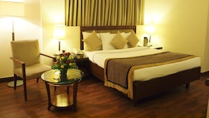 Una habitación de hotel con cama, dos lámparas de noche, una silla, una mesita con un jarrón de flores y un ventanal con cortinas.