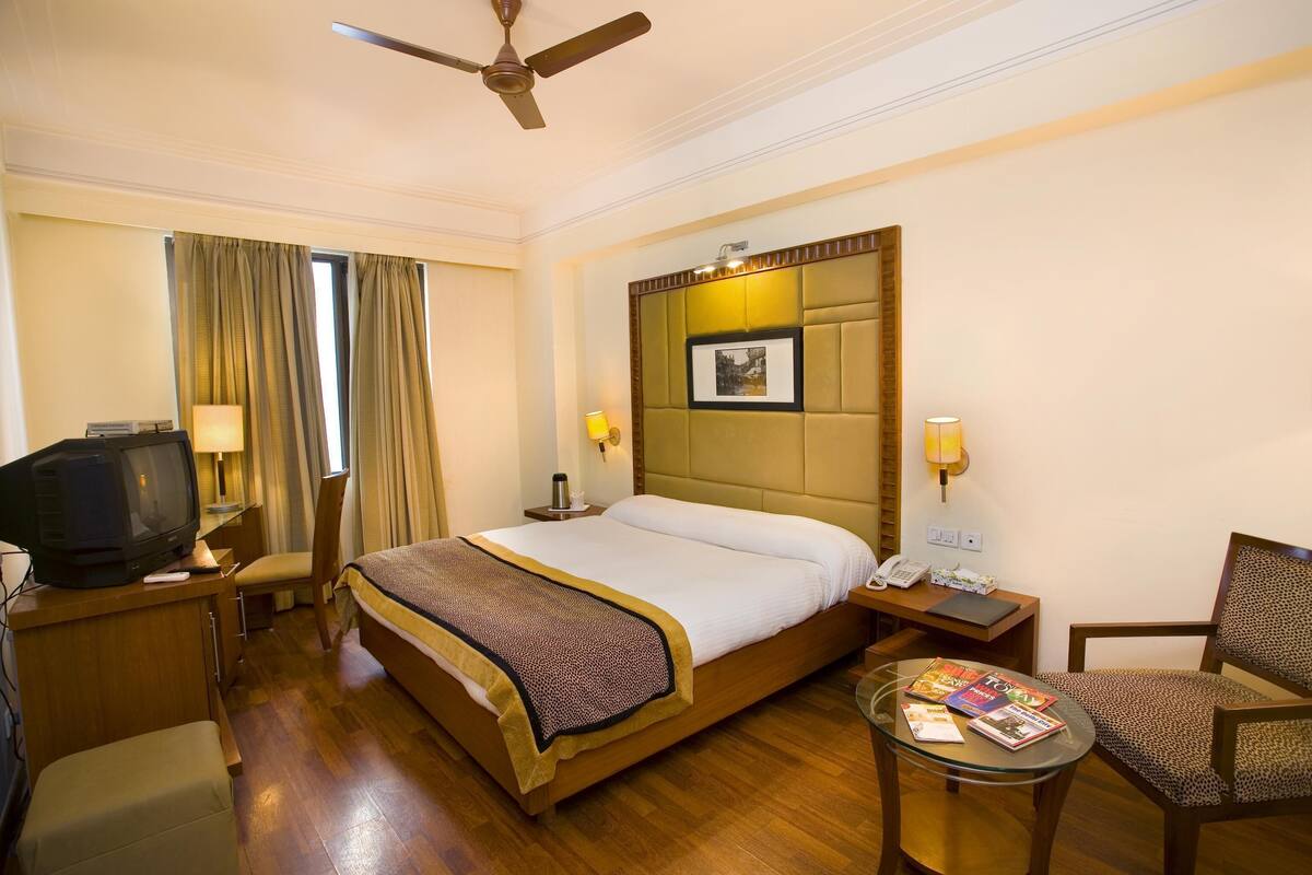 A hotel room with a bed, a television, a desk with a lamp, a chair, and a small table with magazines.
