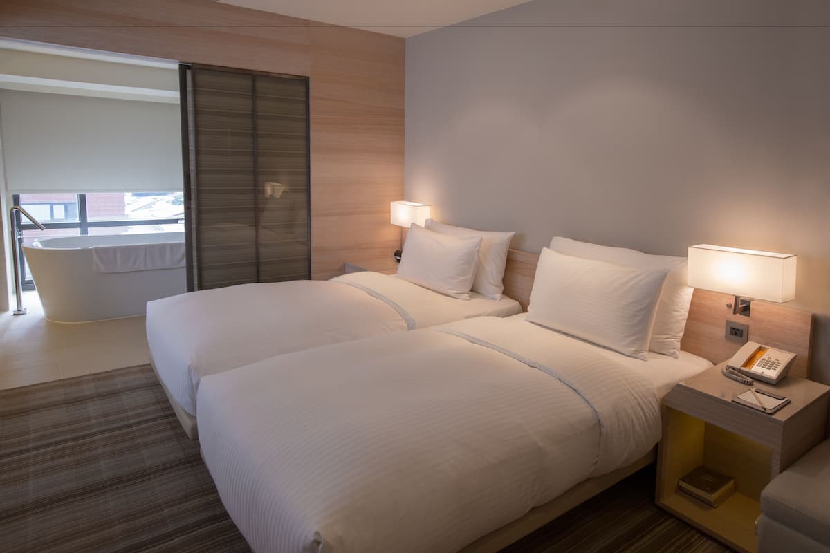 Habitación de hotel con dos camas, una mesita de noche con lámpara, teléfono y vistas a un balcón.