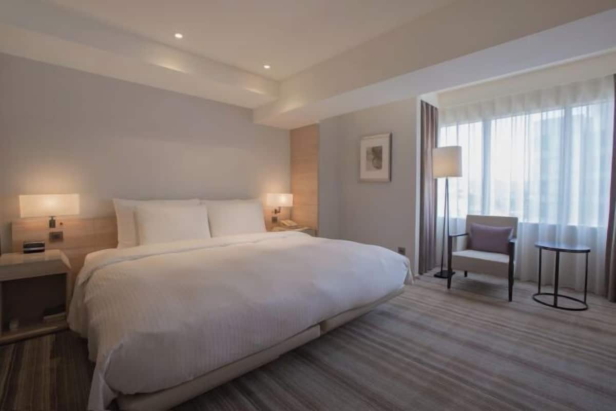 Habitación de hotel con una cama grande, lámparas de noche, una silla, una mesita y una ventana con cortinas.
