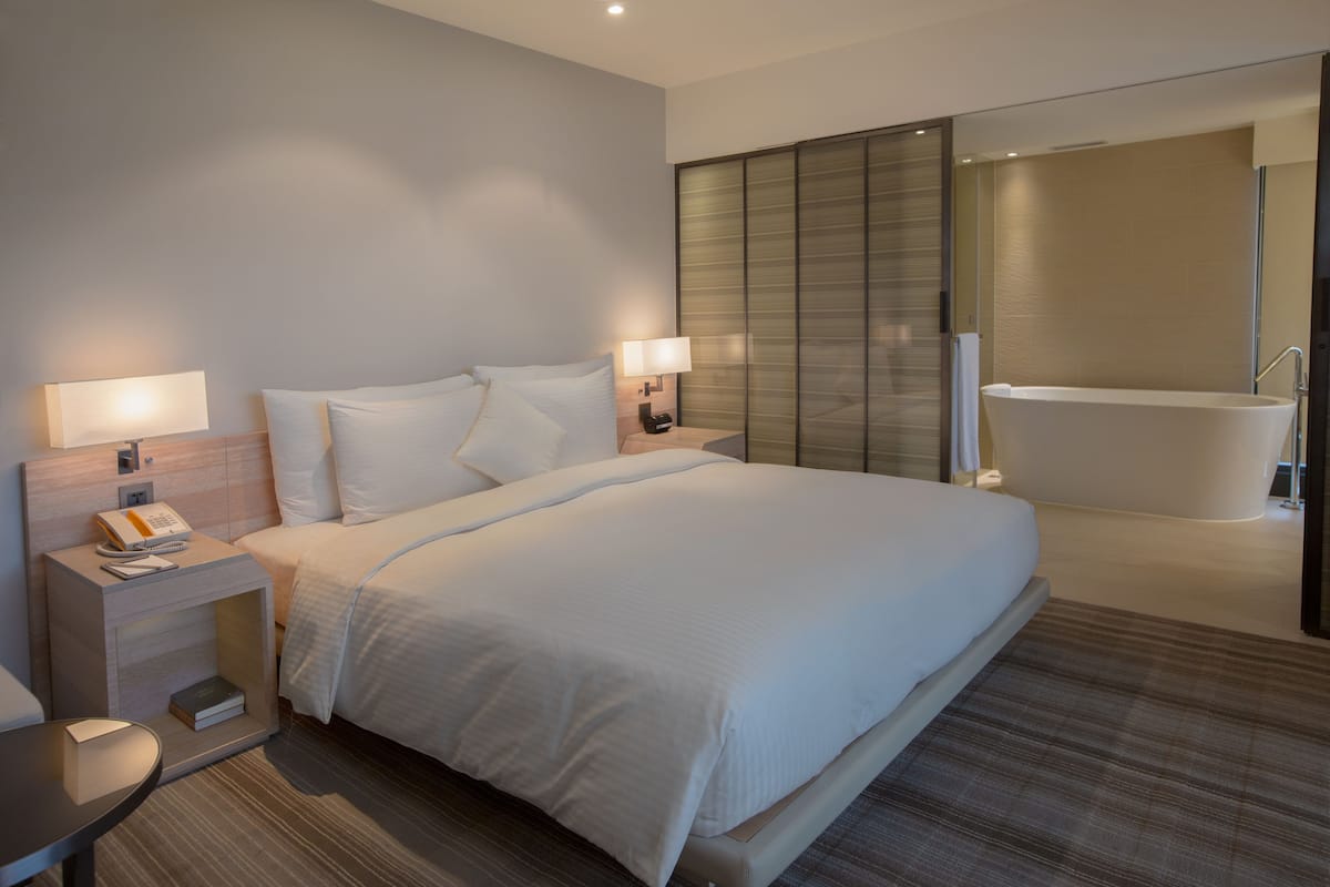 Una habitación de hotel moderna con una cama grande, lámparas de noche, teléfono junto a la cama, una mesita y un baño con bañera.