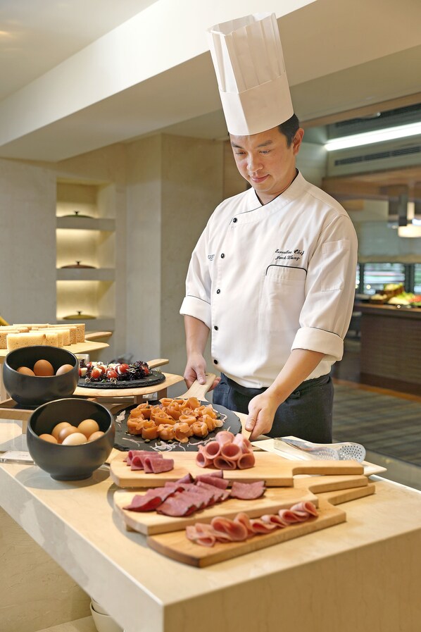Un chef con uniforme blanco y un alto gorro de cocinero, preparando comida en una barra con diversos ingredientes y platos.