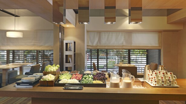Montaje de bufé con diversos alimentos y una estación de café en un interior moderno con detalles de madera y ventanas amplias.