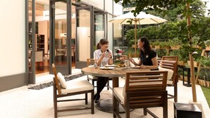 Un área de comedor al aire libre con mesas y sillas de madera, rodeada de follaje y una puerta de vidrio que da a un espacio interior.