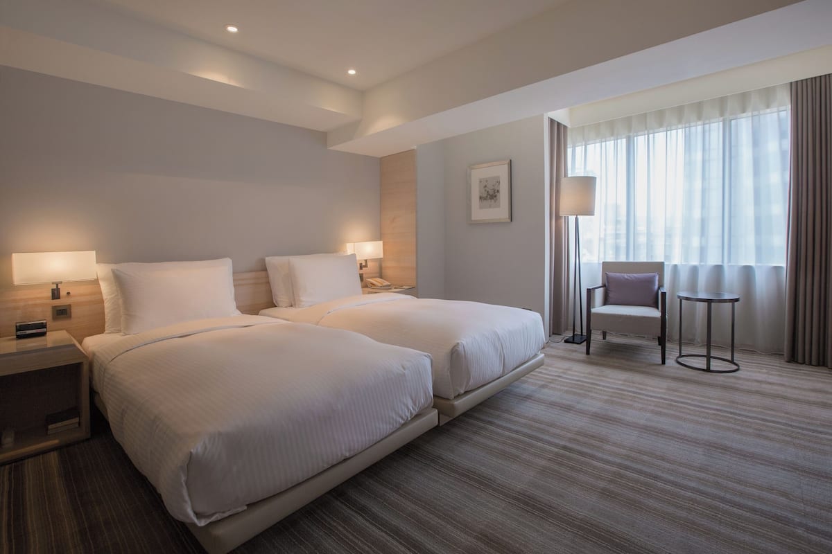Habitación de hotel con dos camas, una silla, una mesita y un amplio ventanal con cortinas.