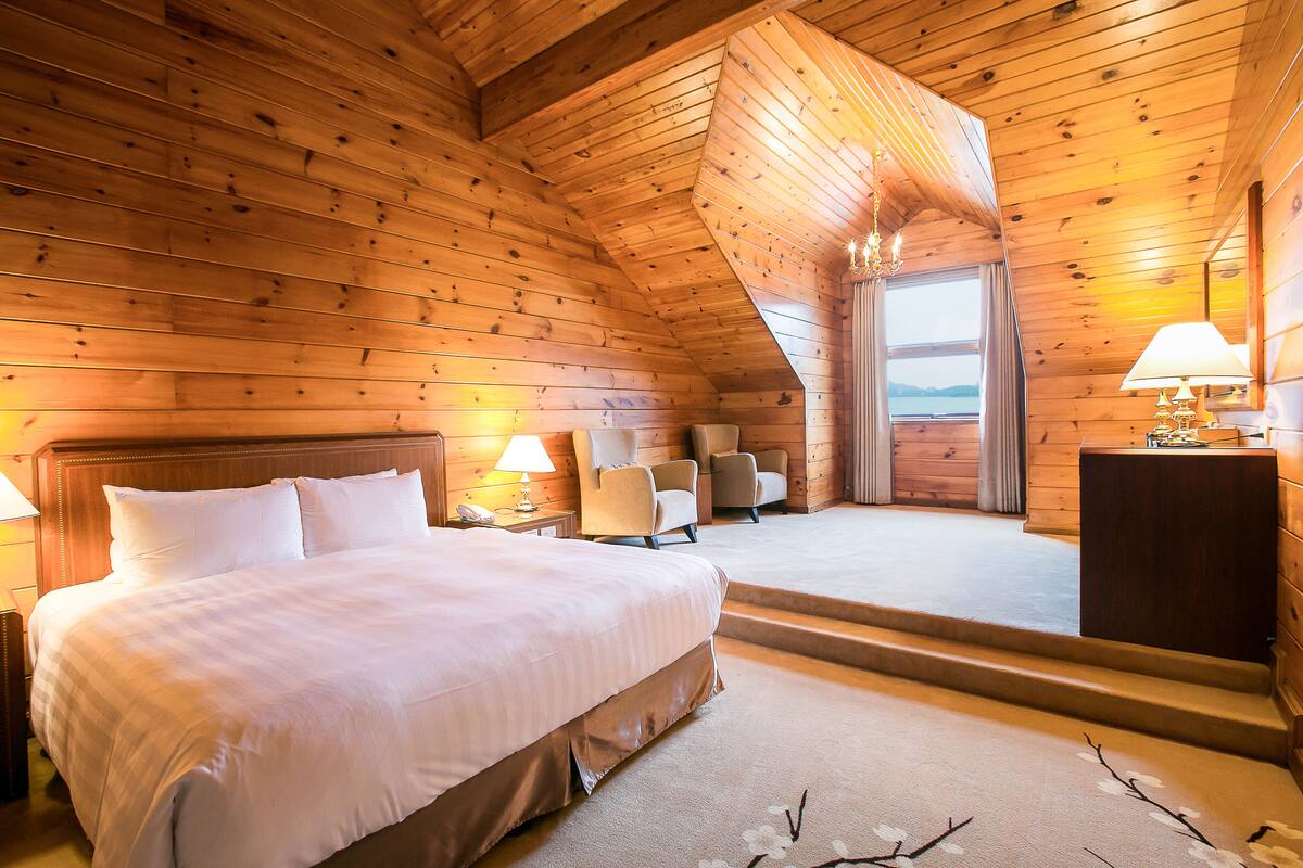 A wooden cabin bedroom with a large bed, two chairs, a desk, and a view of the outdoors.