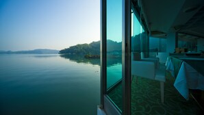A room with a large window overlooking calm water and a distant shoreline. Inside, there is a dining table with chairs and a patterned carpet. - LEALEA GARDEN HOTEL SUN MOON LAKE (Yuchi)