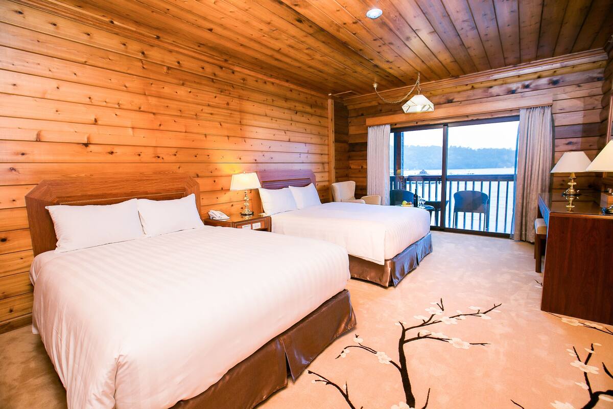 A wooden cabin room with two beds, a balcony, and a view of the outdoors.