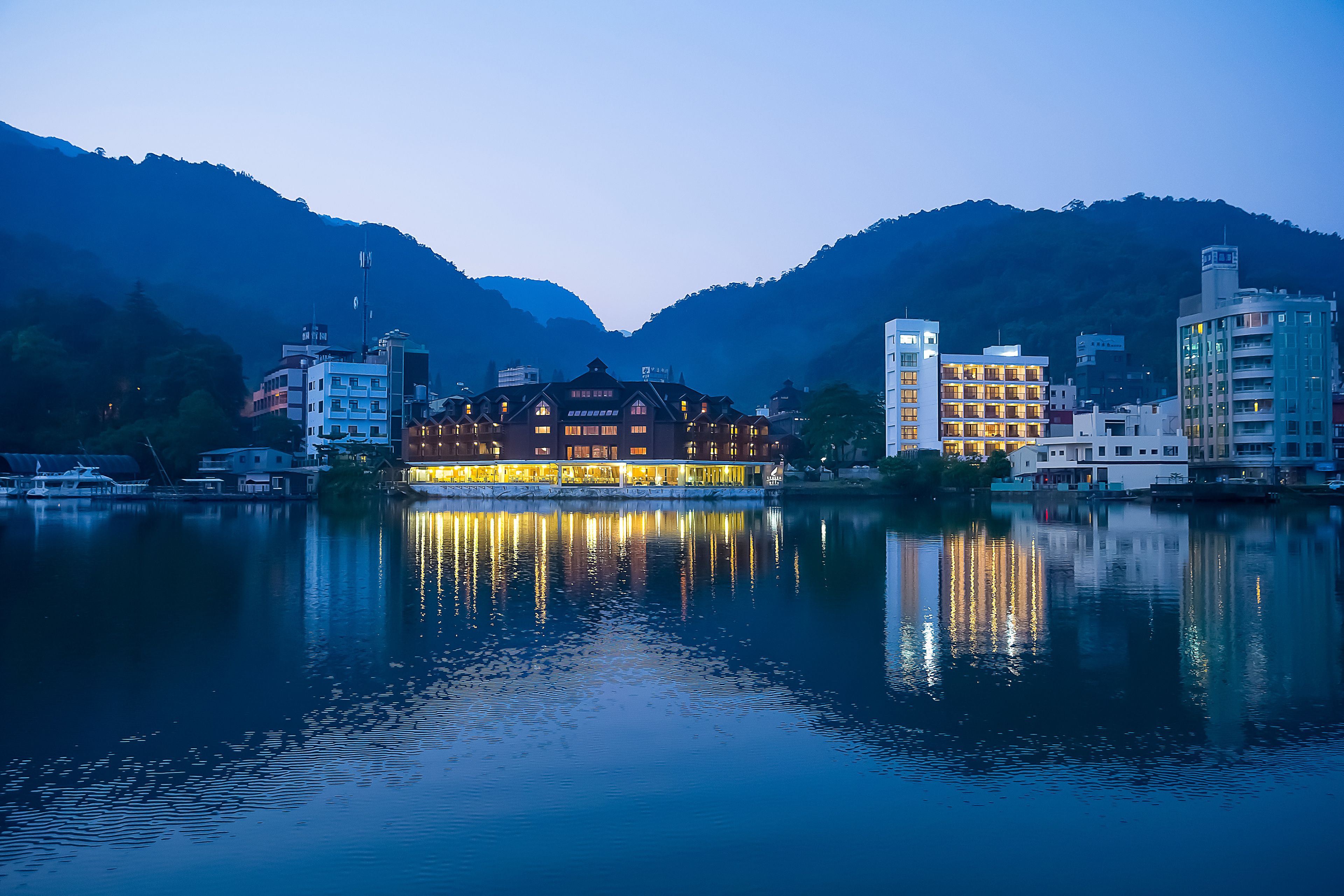 A waterfront hotel with a traditional architectural style, reflecting in calm water, surrounded by mountains.