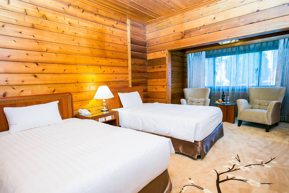 A wooden cabin-style room with two beds, a nightstand with a lamp, a chair, and a small table with a bottle and fruit.