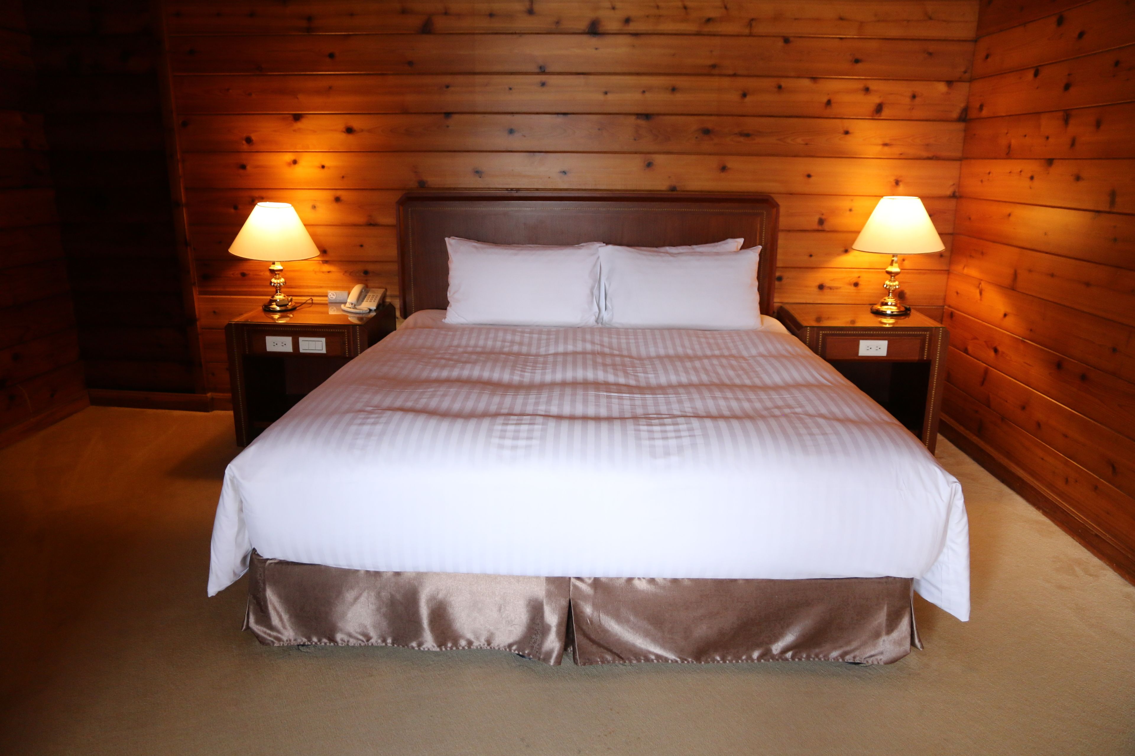 A neatly made bed with a white comforter and pillows, flanked by two bedside tables with lamps, set against a wooden paneled wall.