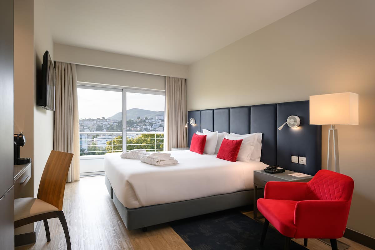 A hotel room with a large bed, a desk, a red chair, a TV, and a view of a cityscape.