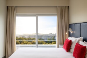 A hotel room with a large window, a bed with white and red pillows, and a nightstand with a lamp.
