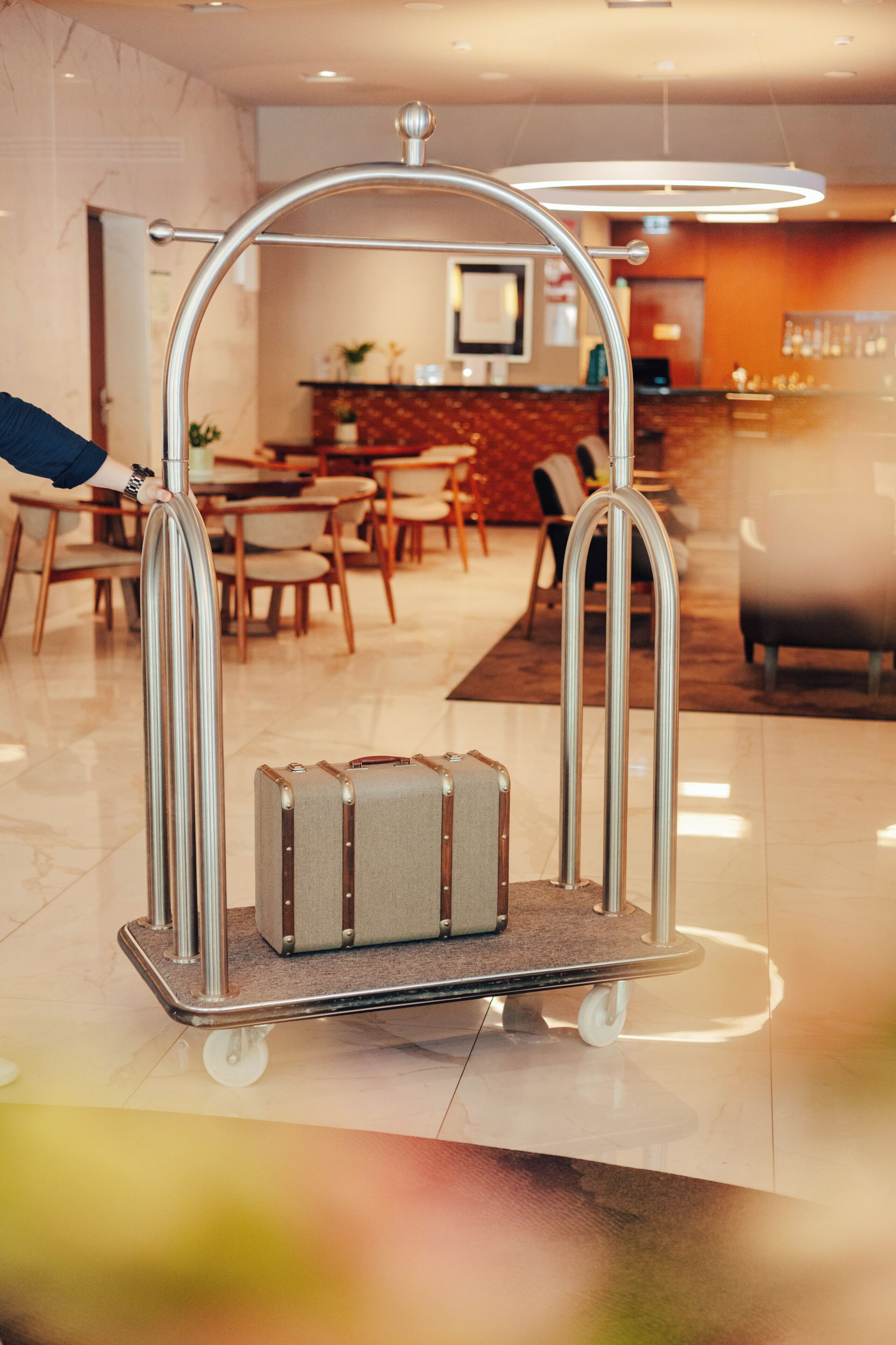 A luggage cart with a suitcase in a hotel lobby with a dining area in the background.