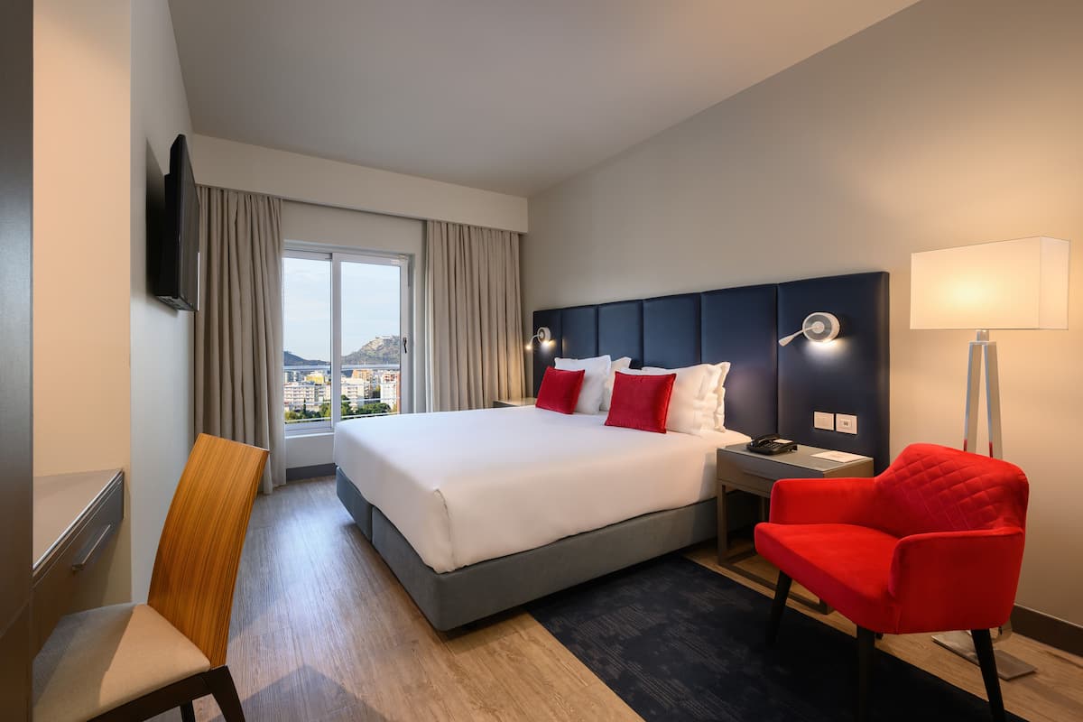 A hotel room with a large bed, a desk, a chair, a TV mounted on the wall, and a view of the city through the window.