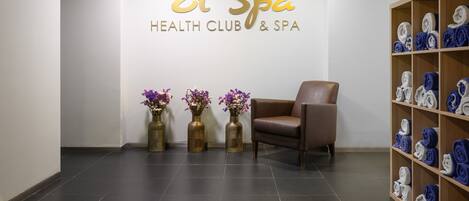 Una zona de recepción con un letrero que dice "El Spa Health Club & Spa", un sillón de cuero marrón y un estante con toallas.