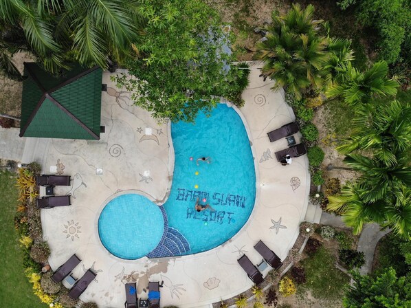 Vue aérienne d’une piscine de forme circulaire, évoquant un soleil, entourée de chaises longues et de palmiers.