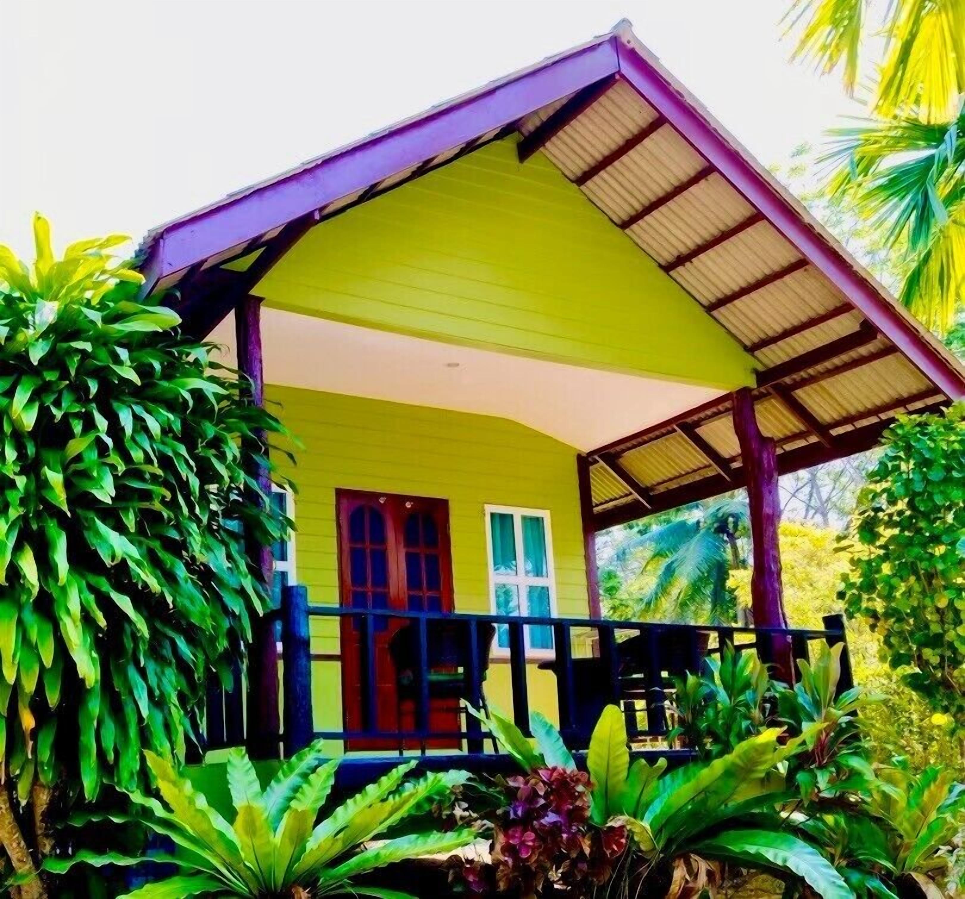 A single-story building with a yellow facade and a purple roof, surrounded by lush greenery.