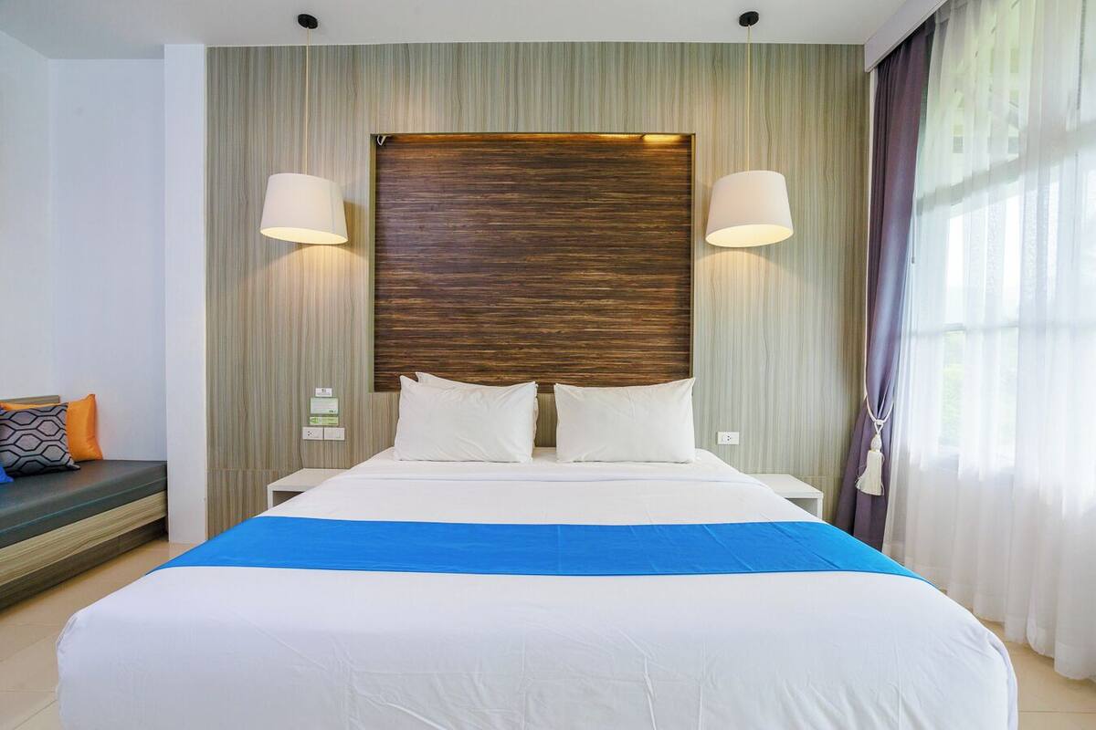 A hotel room with a large bed, two bedside lamps, a headboard with a wood-like texture, a blue accent on the bedspread, and a window with curtains.