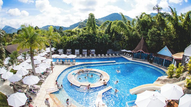 A large outdoor swimming pool with multiple lanes, surrounded by lounge chairs and umbrellas, set against a backdrop of lush greenery and mountains. - Hotel Village Le Canton (Teresópolis)