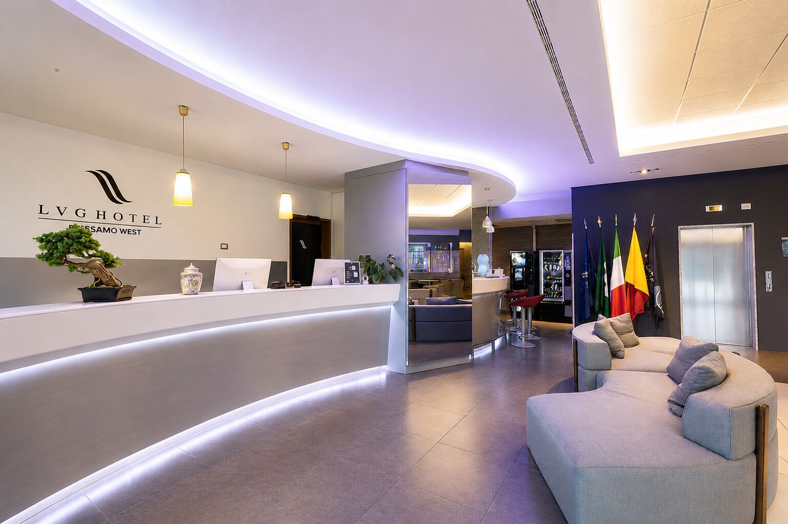 Ein Hotel-Empfangsbereich mit einer gebogenen Rezeption, einer Sitzecke mit einer Couch und einer Bar mit Flaggen.