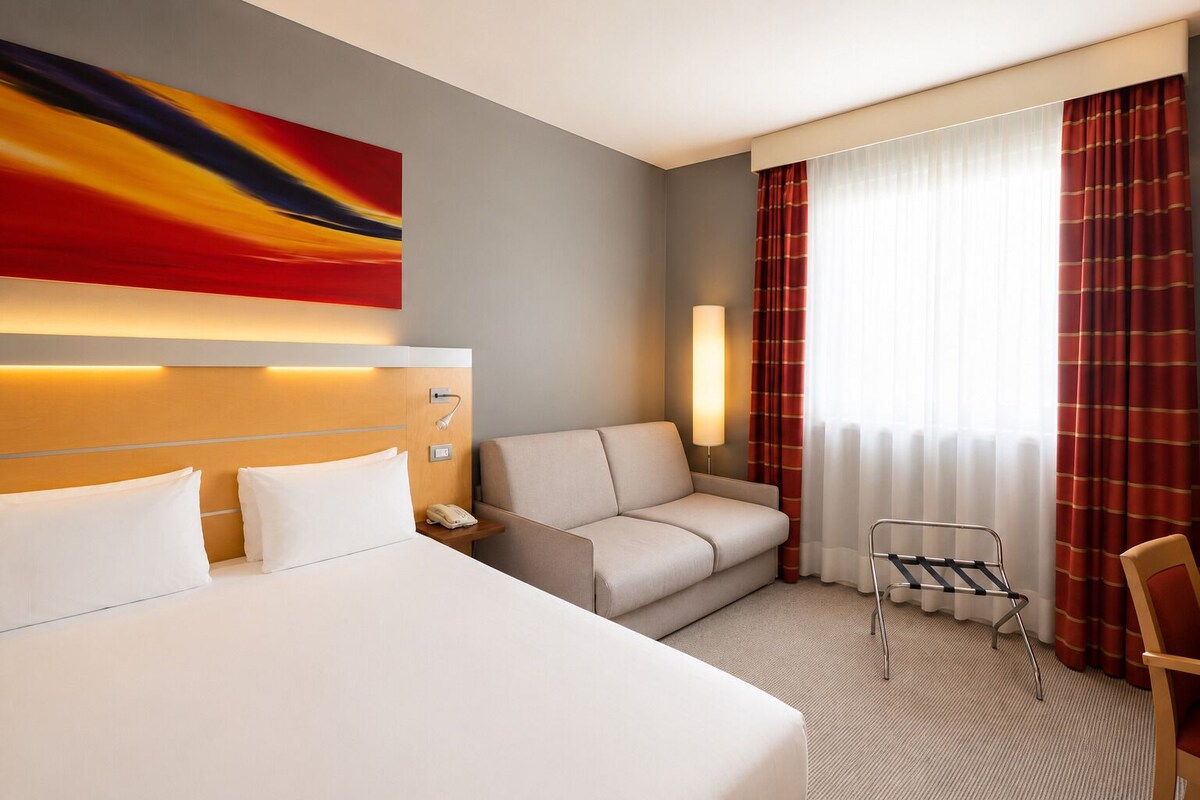 A hotel room with a bed, a sofa, a desk, a chair, a lamp, and a painting on the wall.