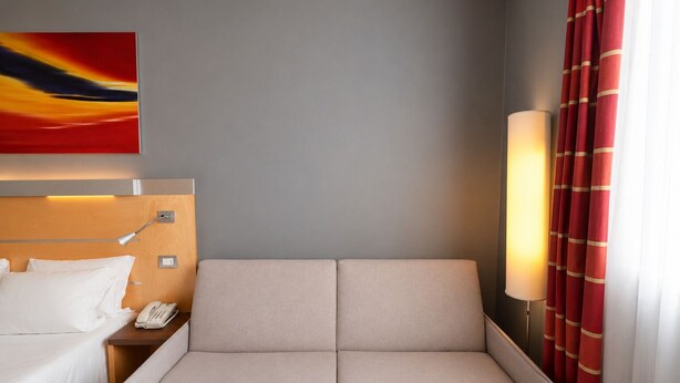 A hotel room with a bed, bedside table, lamp, and a painting on the wall. - LVG Hotel Collection - Bergamo West (Mozzo)