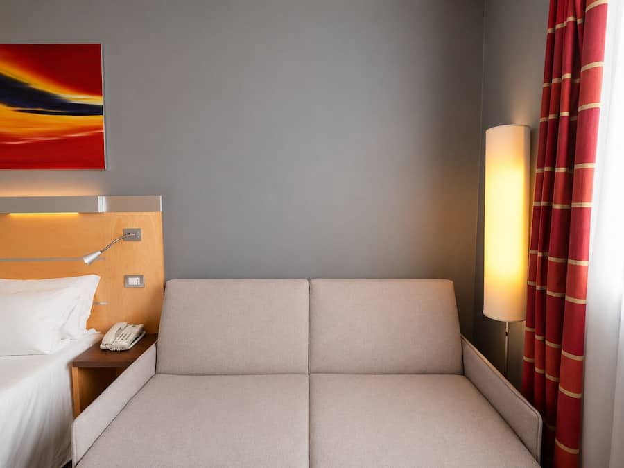 A hotel room with a bed, bedside table, lamp, and a painting on the wall.