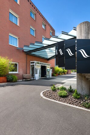 Entrada de hotel con una cubierta de vidrio, un banco y un camino que lleva a la entrada.