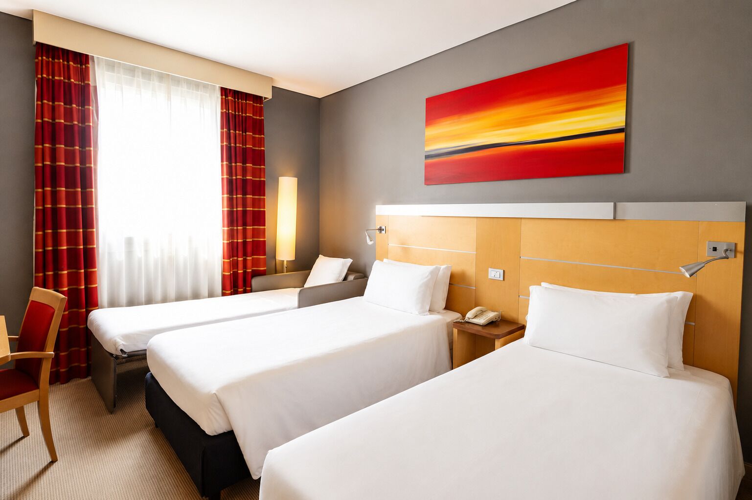 Ein Hotelzimmer mit drei Betten, einem Schreibtisch, einem Stuhl, einer Lampe und einem Bild an der Wand.
