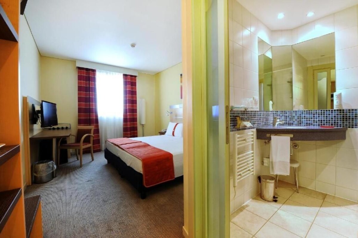 A hotel room with a bed, desk, chair, and a bathroom with a mirror, sink, and towel rack.