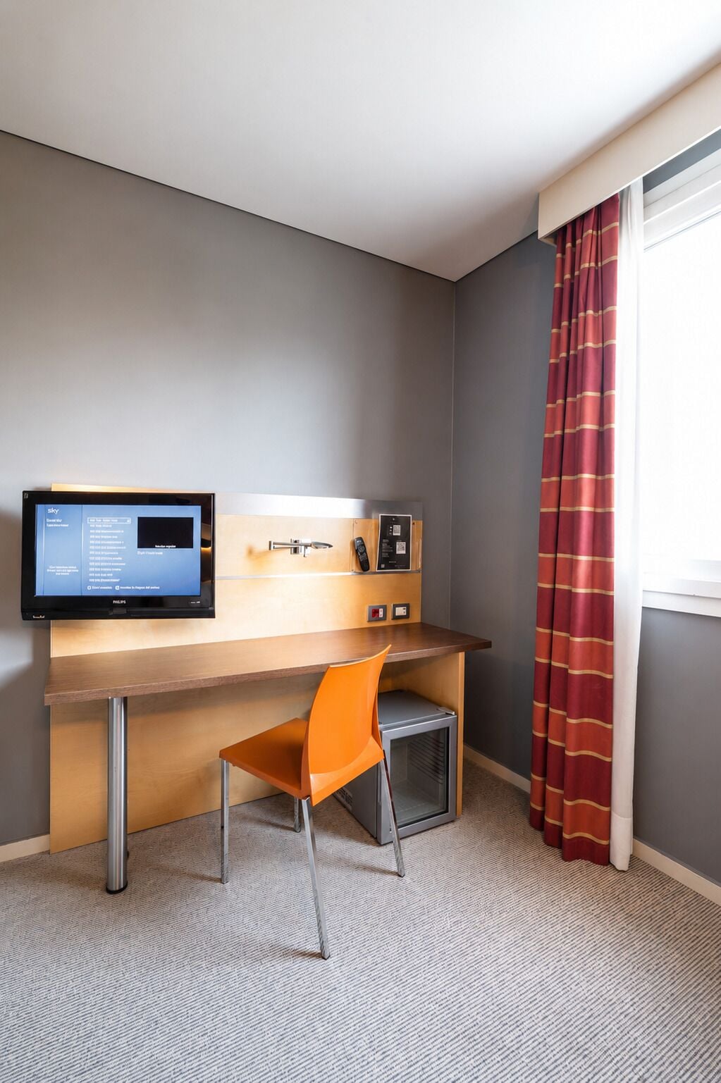 Habitación de hotel con escritorio, silla, televisor, mini refrigerador y ventana con cortinas.