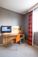 Habitación de hotel con escritorio, silla, televisor, mini refrigerador y ventana con cortinas.