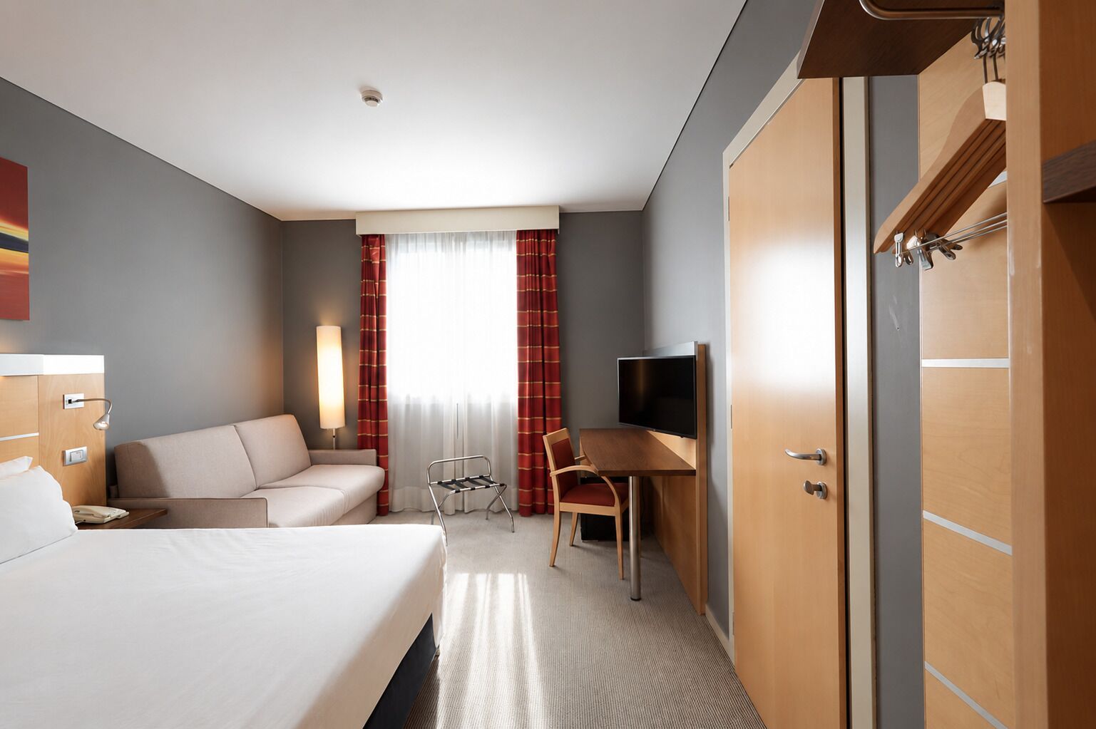 A hotel room with a bed, a desk with a chair, a TV, and a wardrobe.