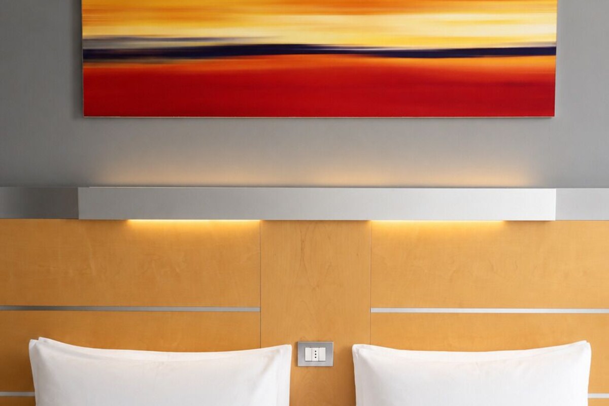A hotel room with a bed, white pillows, a red and orange abstract painting above the bed, and a wooden headboard with a light switch.