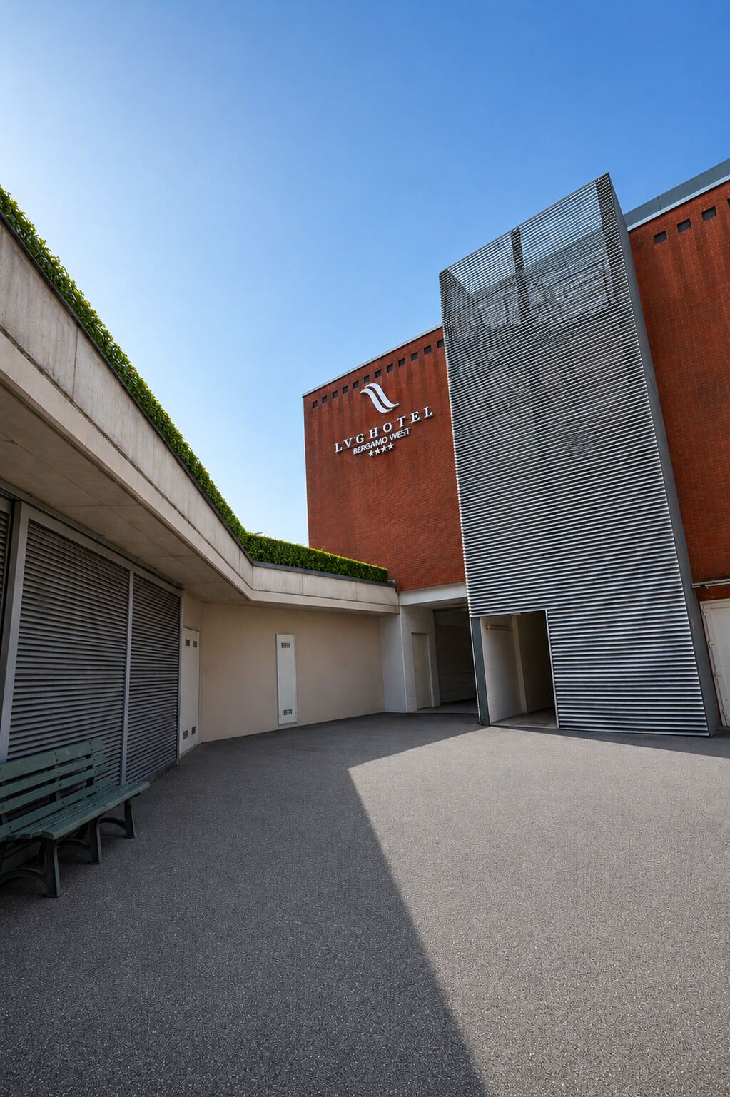 Ein modernes Gebäude mit einem Schild für 'LVGHOTEL BERGAMO WEST' und einer überdachten Außenbereits mit einer Bank.
